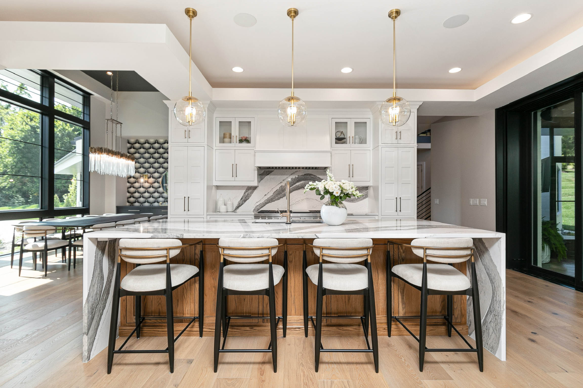 A kitchen counter with four white barstools, three pendant lights, white cabinets, and a marble backsplash